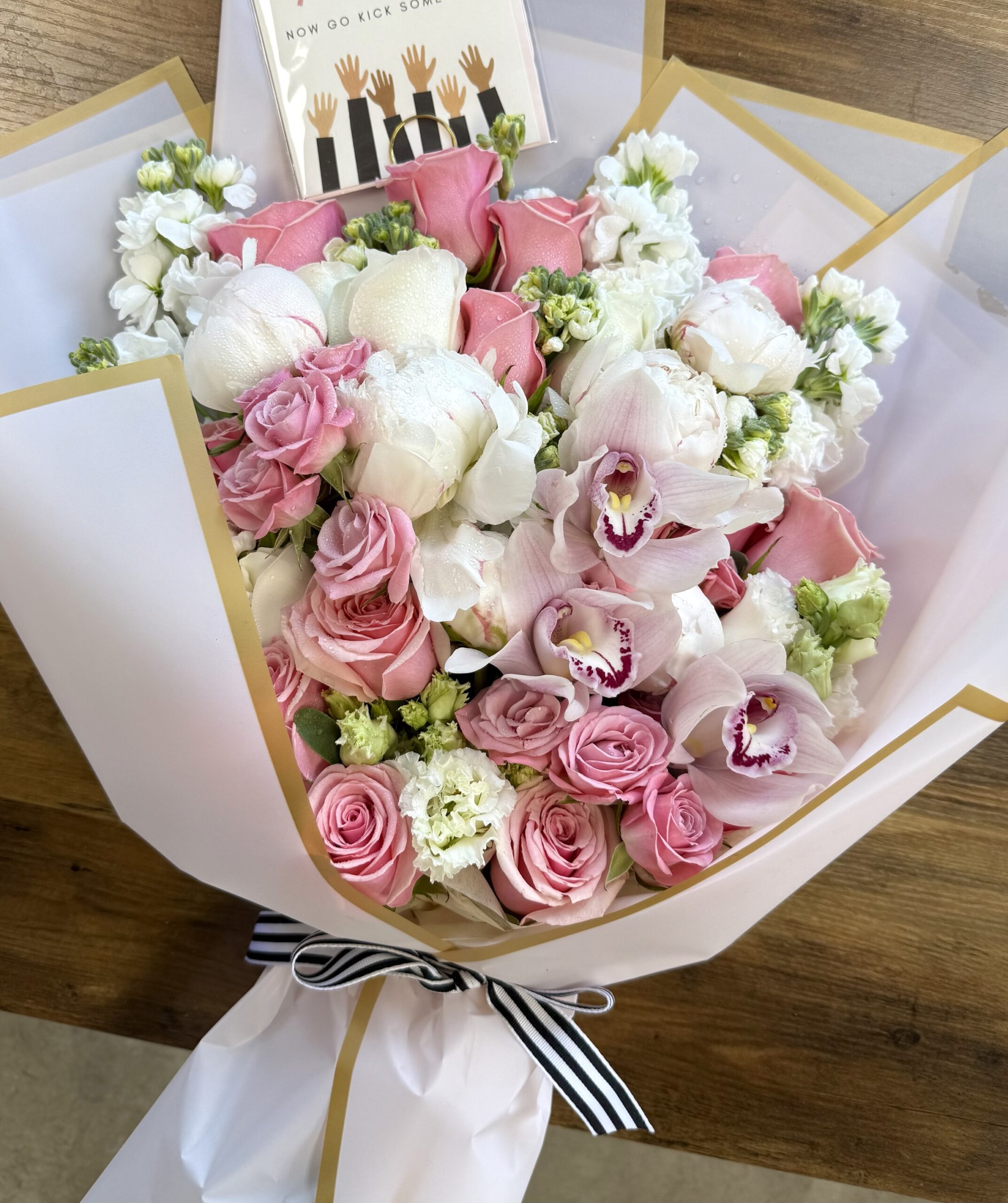 The Skip A Beat - Wrapped Bouquet from Orange County Flowers features pink and white roses, white orchids, and small white flowers, elegantly wrapped in white paper with a black and white ribbon and a card included.