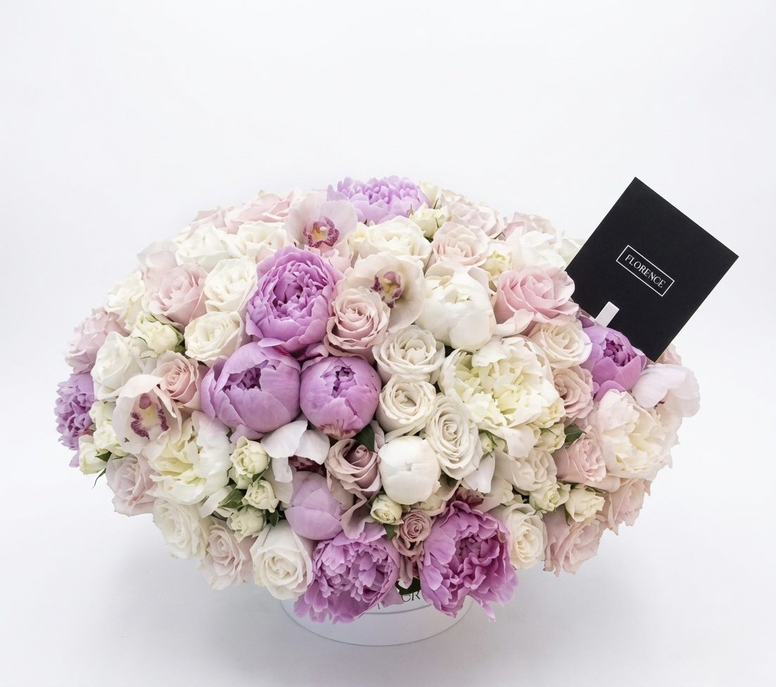 The Legend - Flower Box from Orange County Flowers features pale pink, white, and lavender peonies and roses in a round white box with a black envelope nestled among the blooms, set against a white background.