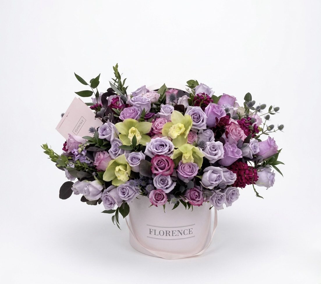 The Violets Are Blue - Flower Box from Orange County Flowers features a white hat box filled with lavender roses, yellow-green orchids, dark purple flowers, and a greeting card, all set against a white background.