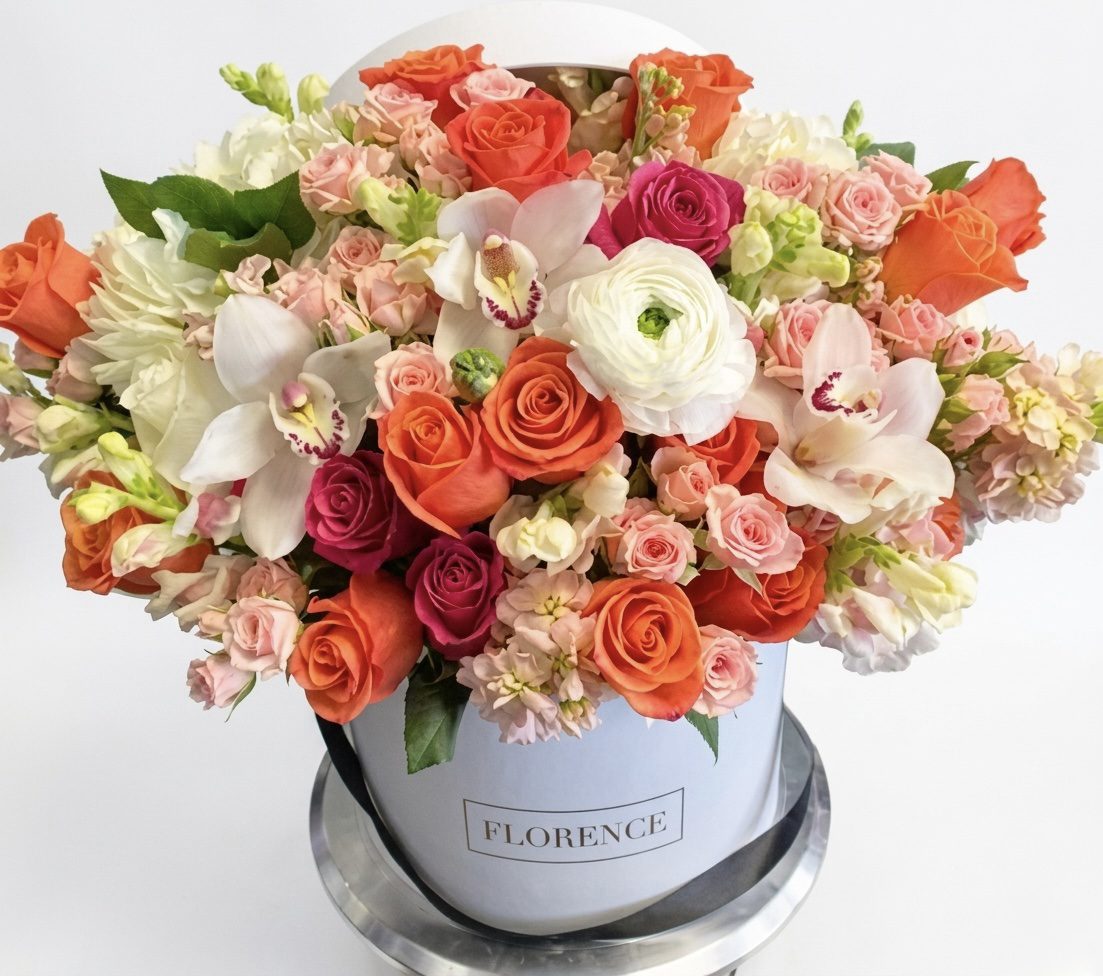 The FLEUR Collection #80 features a large white hatbox filled with orange roses, pale pink spray roses, white ranunculus, and white orchids with pink centers, all elegantly arranged on a round silver tray.