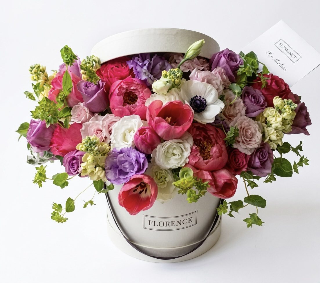 The FLEUR Collection #84 features a round white hatbox filled with pink, purple, white, and red flowers from Orange County Flowers. A small card is nestled among the vibrant blooms.