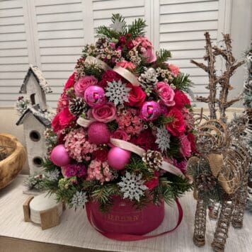 The Very Pink Xmas - Flower Box is a festive Christmas tree-shaped floral arrangement with pink and red flowers, pink ornaments, snowflake accents, and greenery in a pink hatbox, surrounded by rustic winter decor on a white table.