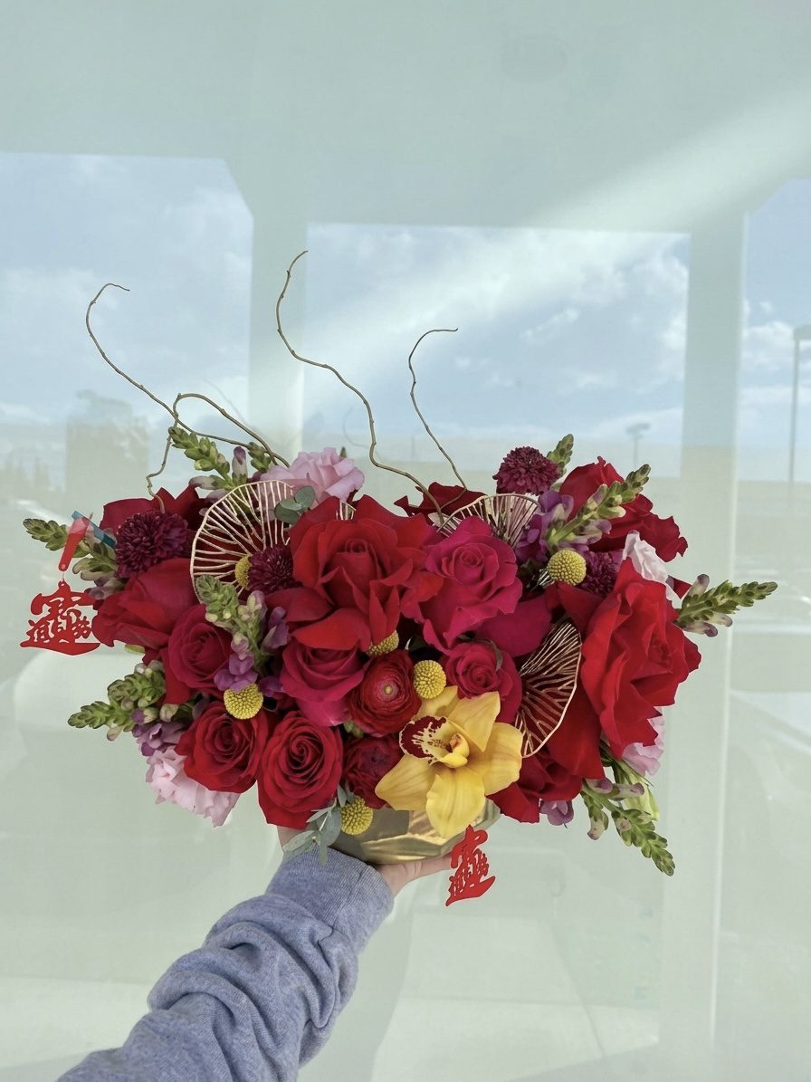 A hand holds the Happy Lunar - Ceramic Pot featuring a vibrant arrangement of red and pink roses, yellow orchids, and gold fans, set against a light background with reflections of the sky and windows.