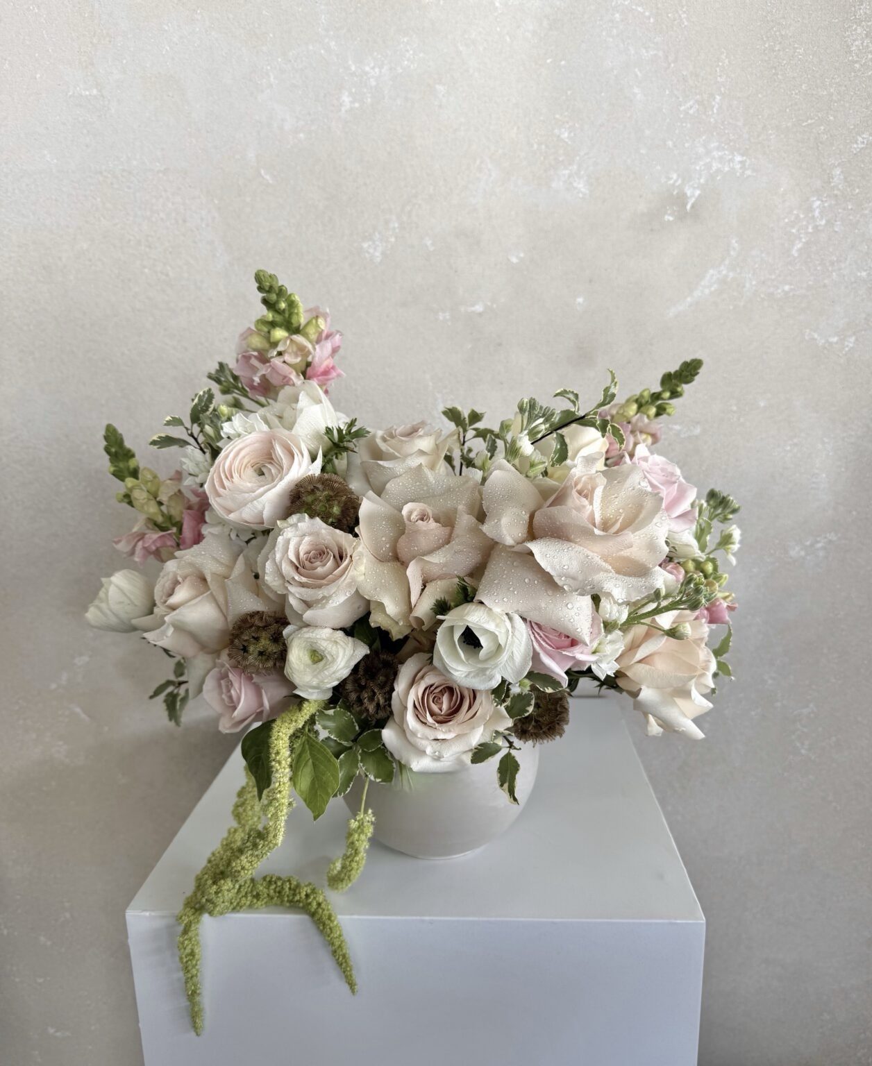 The Posh - Ceramic Pot displays an elegant mix of pale pink and cream roses, lisianthus, snapdragons, and greenery, arranged against a light textured wall on a white pedestal.