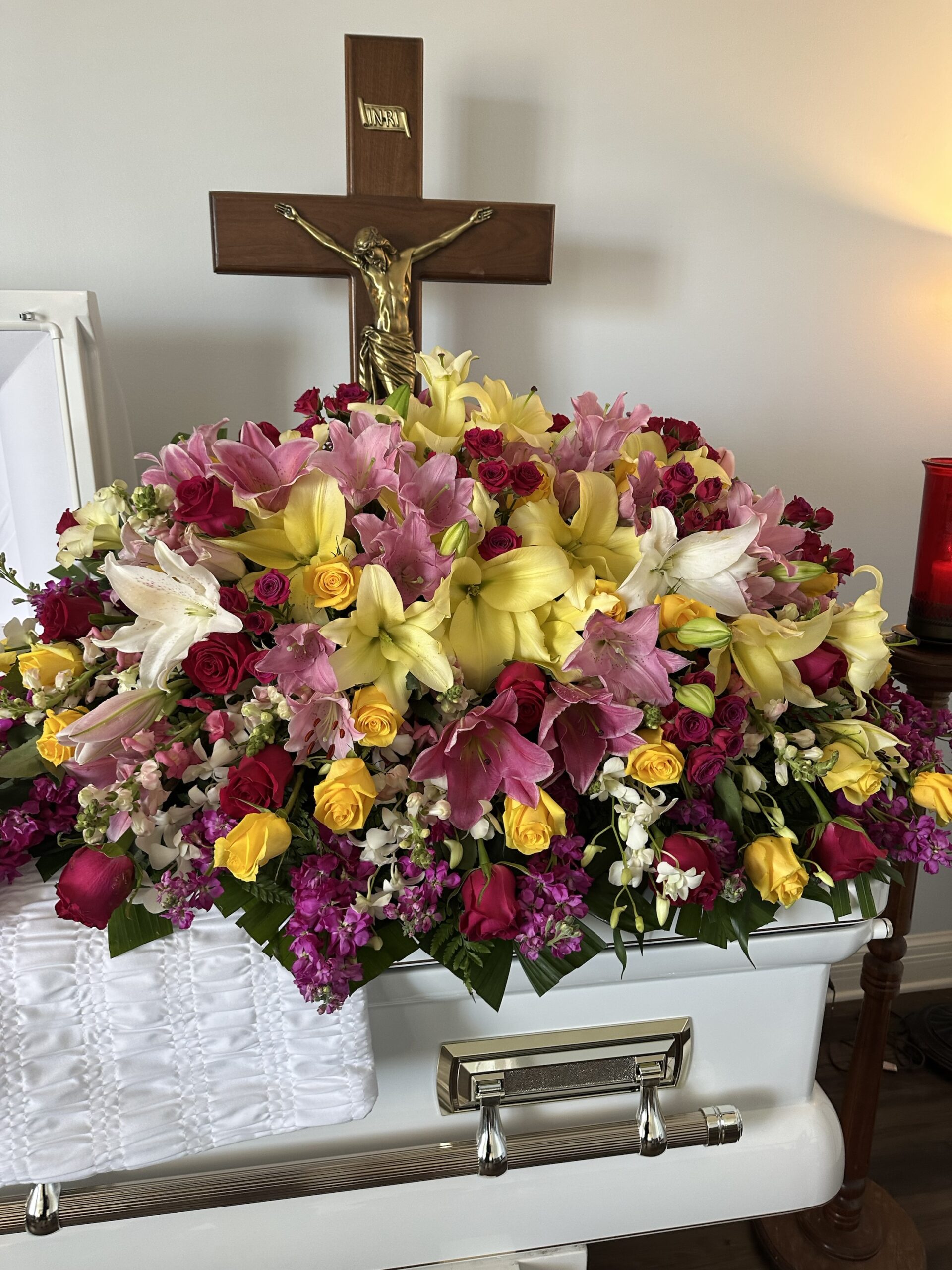 The Full of Life - Casket, a white casket topped with a vibrant floral arrangement of lilies and roses, rests beneath a crucifix in a softly lit room.