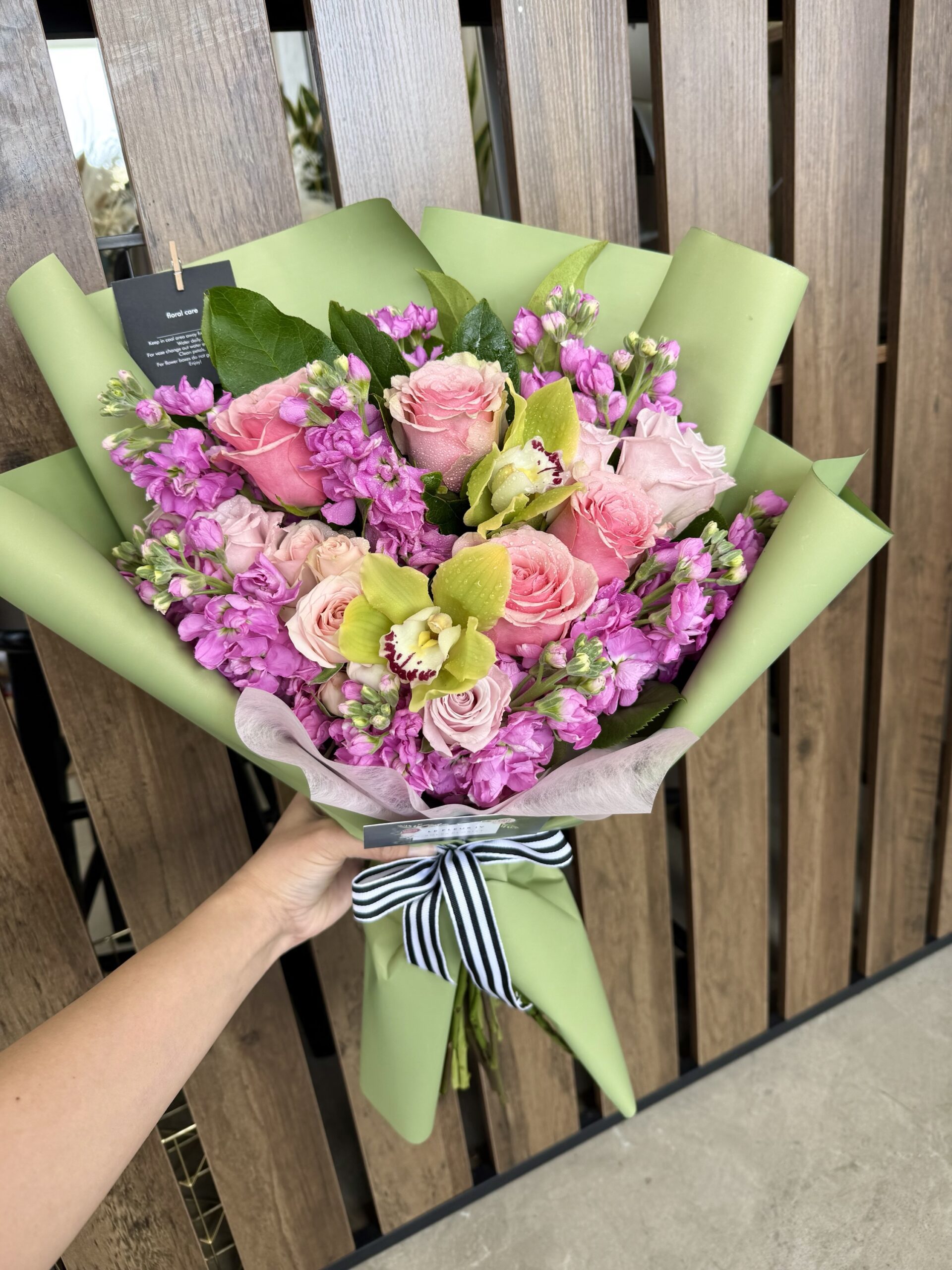 A hand holds the Pistachio Rose - Wrapped Bouquet, featuring pink roses, purple blooms, and green orchids in light green paper with a black-and-white striped ribbon, set against a wooden slat background.