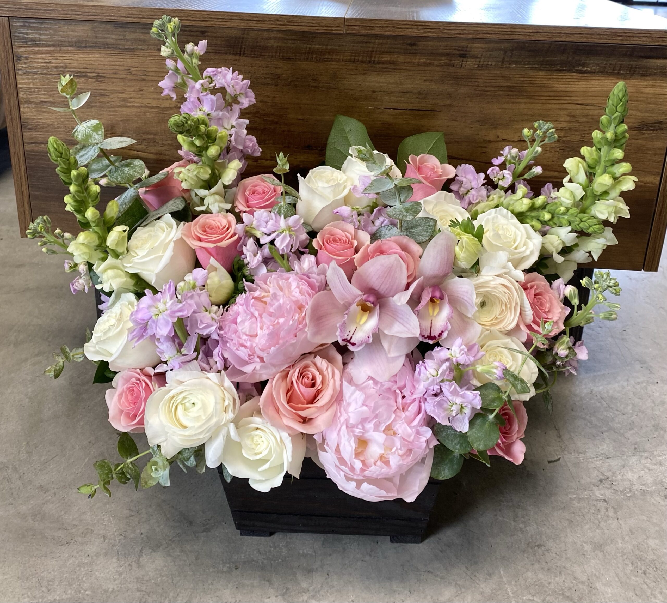 The Socialite - Wood Pot is a rectangular black box featuring a lush mix of pink and white roses, peonies, orchids, and greenery, displayed on a gray floor in front of a wooden table.