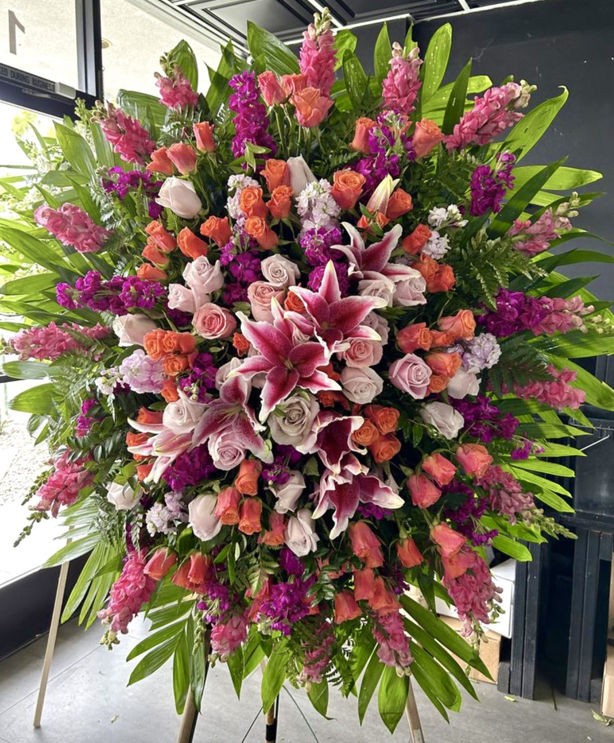 The Celebration Of Life - Funeral Spray is a large, colorful arrangement of pink lilies, pink and peach roses, purple blooms, and lush greenery, elegantly displayed on an indoor stand.