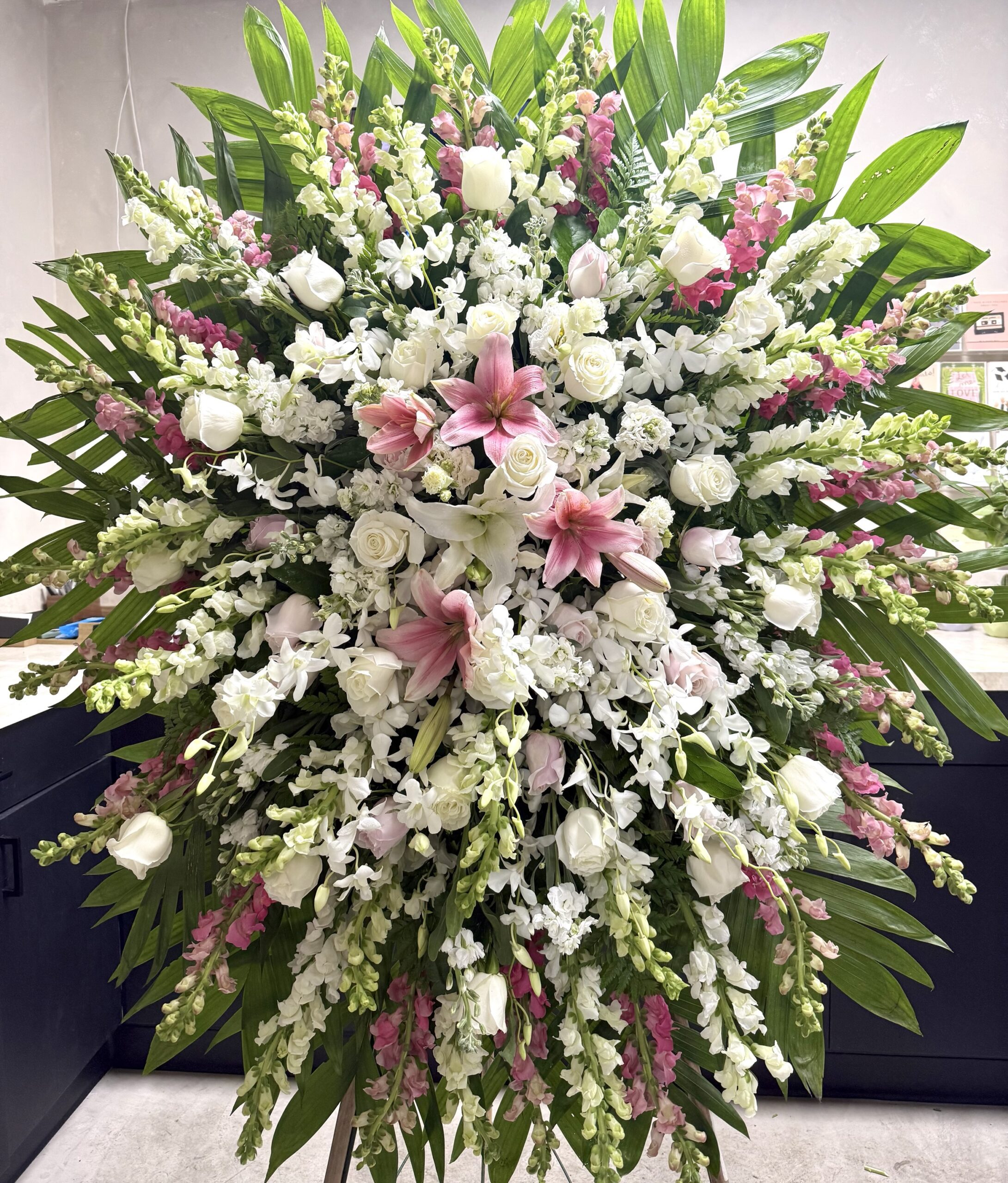 The Into The Light - Funeral Spray features a large floral arrangement of white roses, lilies, orchids, snapdragons, pink lilies, and green leaves, beautifully displayed against a plain indoor background.