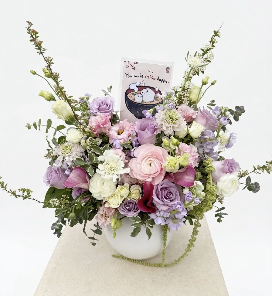 The Bella - Ceramic white vase displays a pastel floral mix of pink, purple, and white flowers. Among the blooms is a card featuring cartoon animals in a miso soup bowl with the message: "You make miso happy.