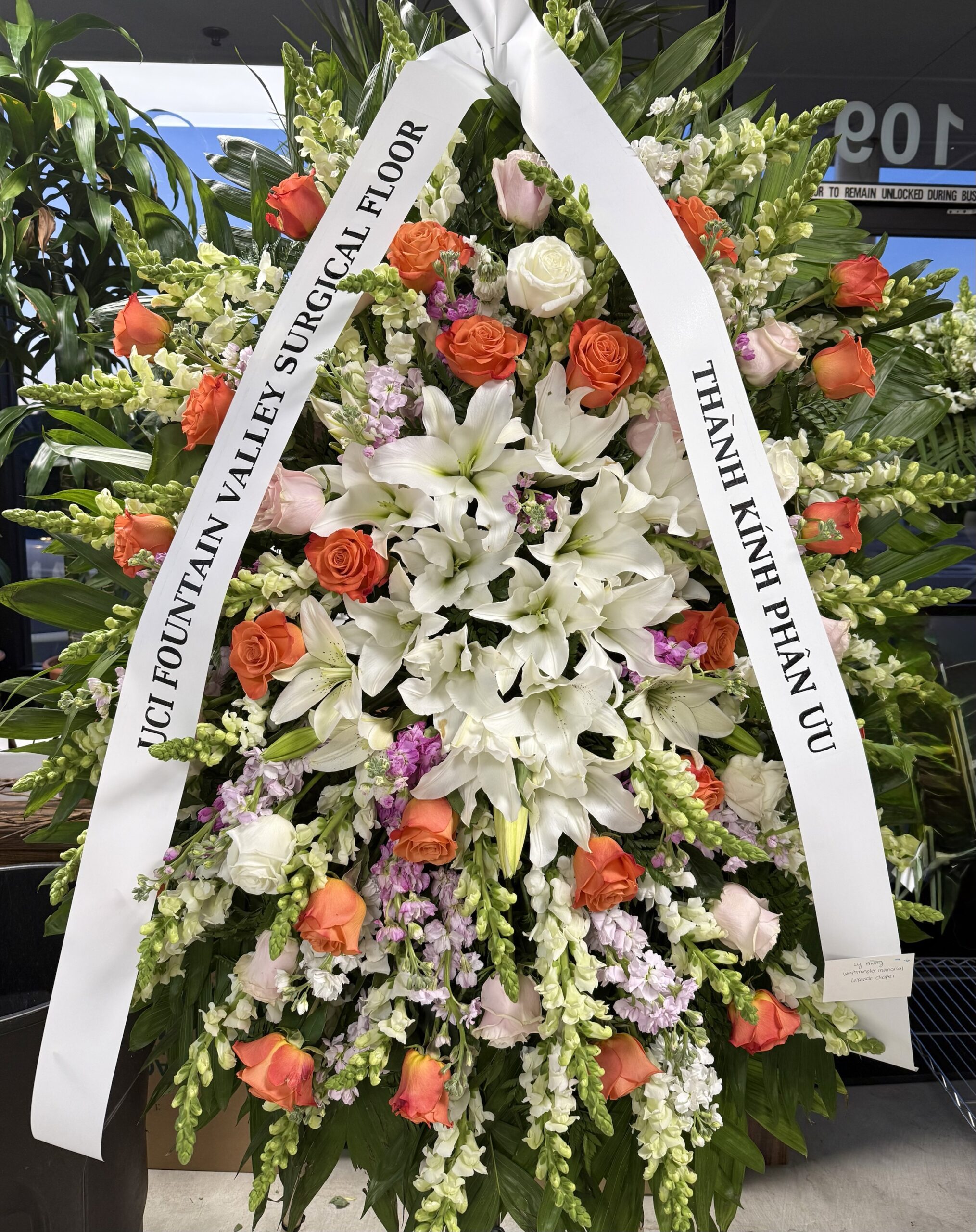 The Eternal Embrace - Funeral Spray features white lilies, orange and pink roses, and greenery. Two white ribbons include condolence messages in English and Vietnamese.