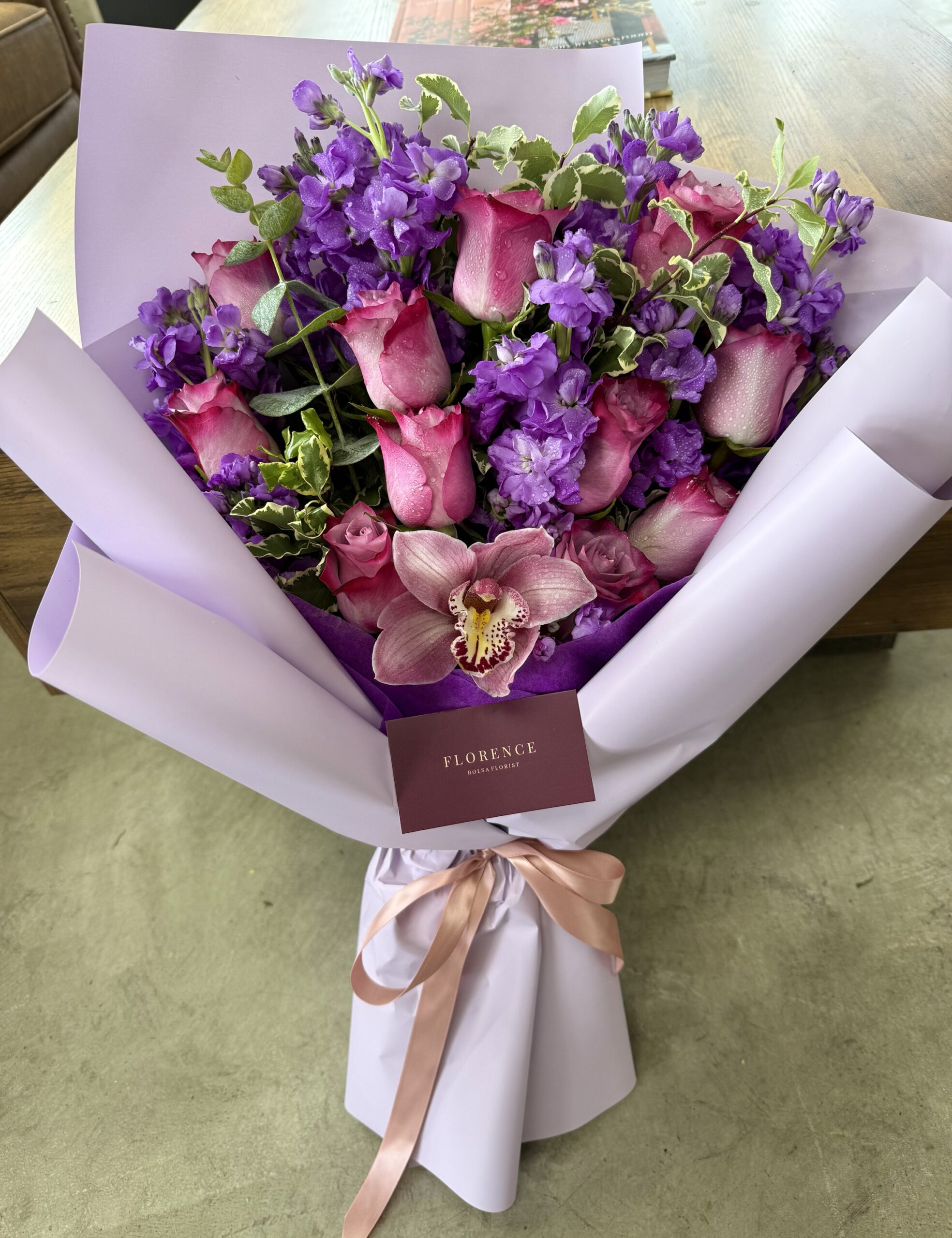 The Dolce Viola - Wrapped Bouquet features pink roses, purple flowers, and a single orchid, elegantly wrapped in pale lavender paper with a pink ribbon and a FLORENCE card.