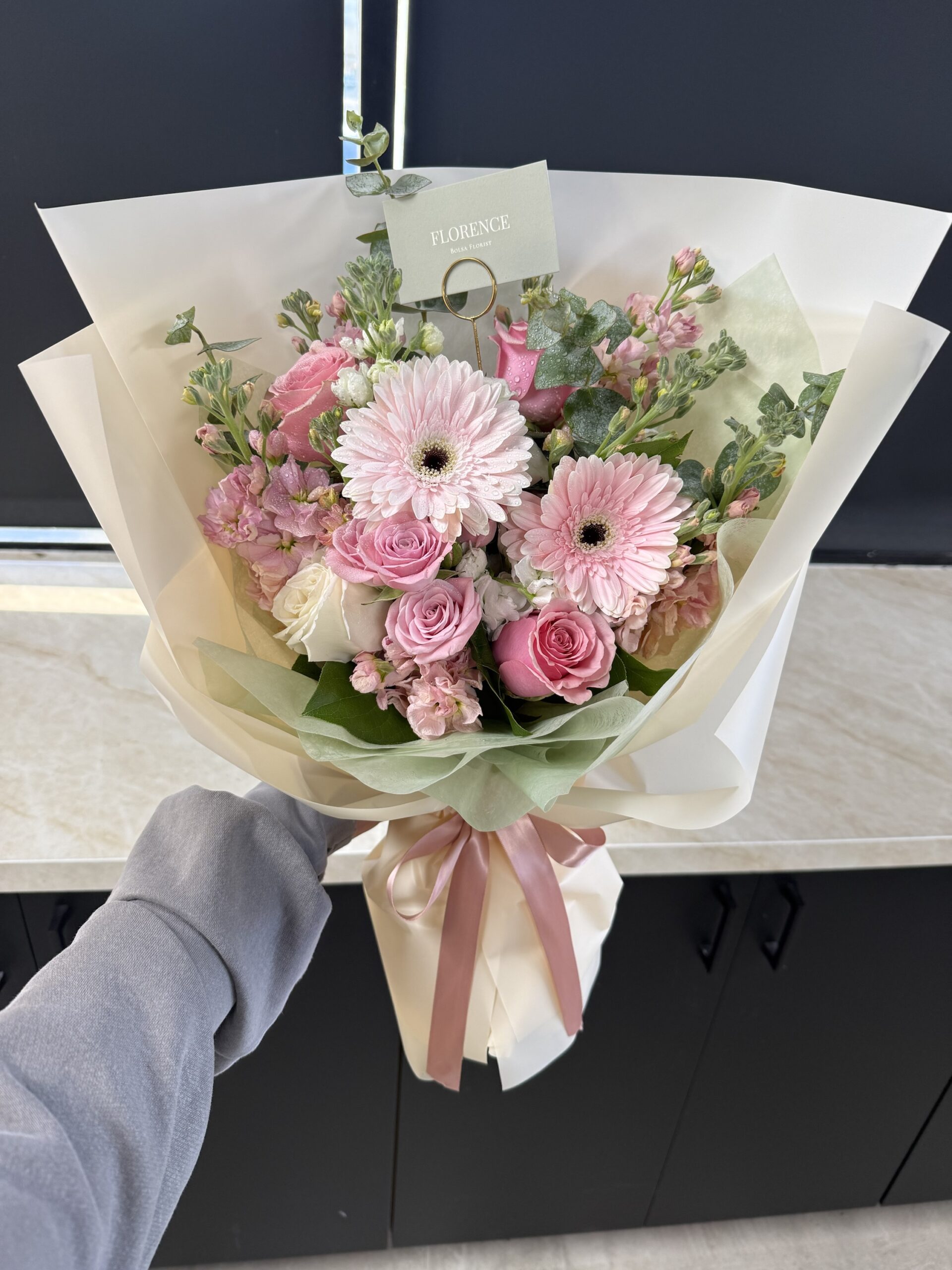 A hand holds the Daisy Me Rollin' - Wrapped Bouquet with pink and white roses and gerberas, wrapped in cream paper and tied with a ribbon. A card labeled “FLORENCE” is tucked among the flowers.