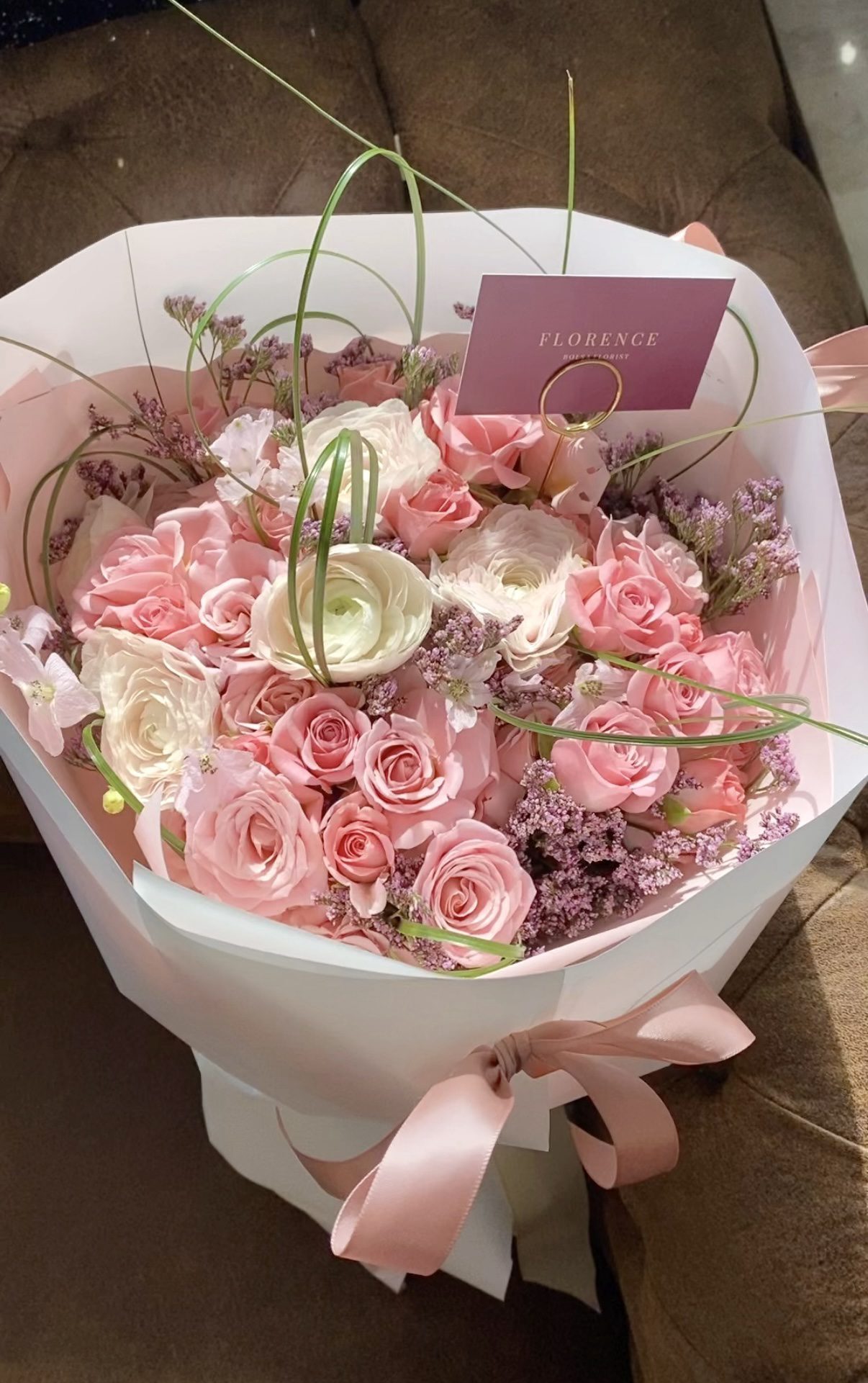The Flirt Alert - Round Wrap Bouquet features pink and white roses with light purple flowers and greenery, elegantly wrapped in white paper with a pink ribbon and a “Florence” card, displayed on a brown surface.