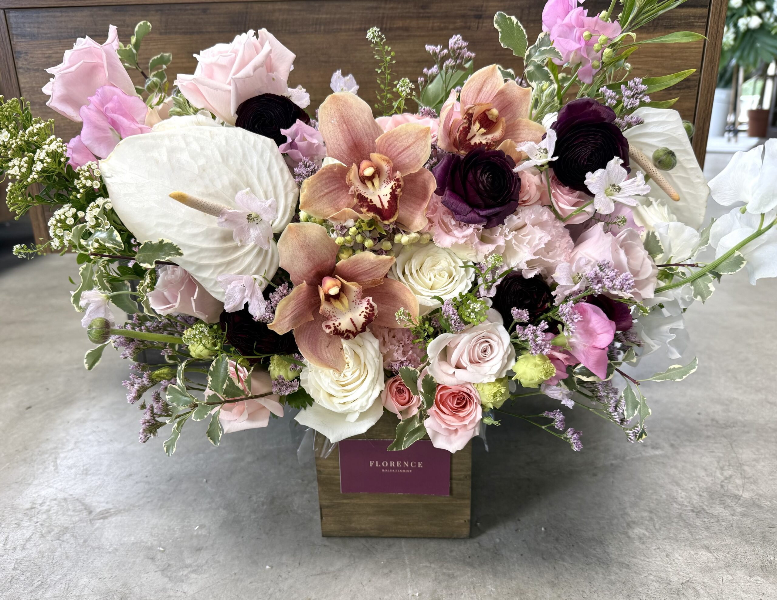 The Rebel Rouge - Wood Pot features a lush floral arrangement with pink roses, pale purple orchids, white anthuriums, dark ranunculus, and greenery in a wooden box displayed on a gray surface.