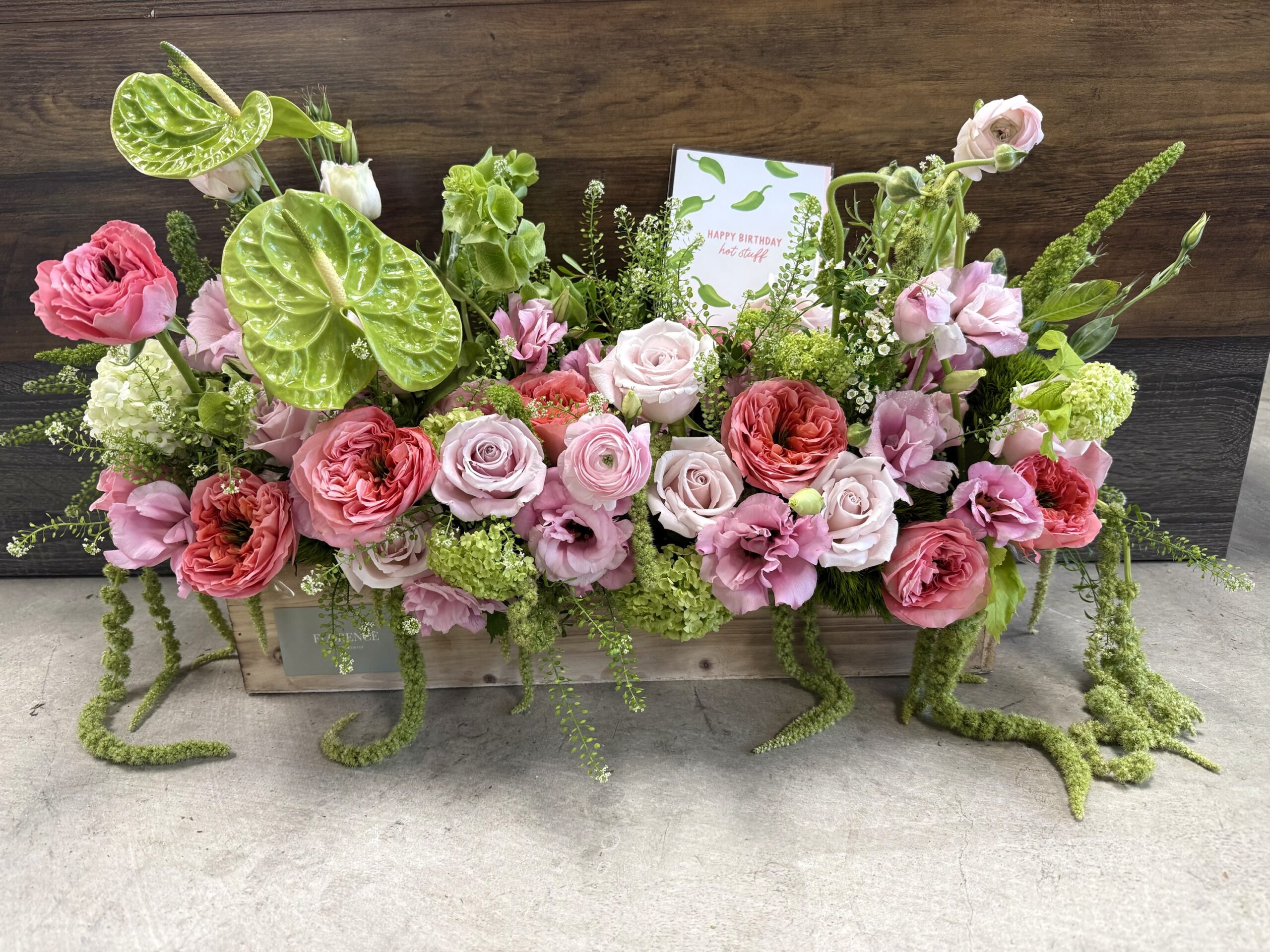 The Mademoiselle - Wood Pot features a lush arrangement of pink and peach roses, pale green anthuriums, and trailing amaranthus in a wooden box, with a "Happy Birthday! We love you!" card among the flowers.