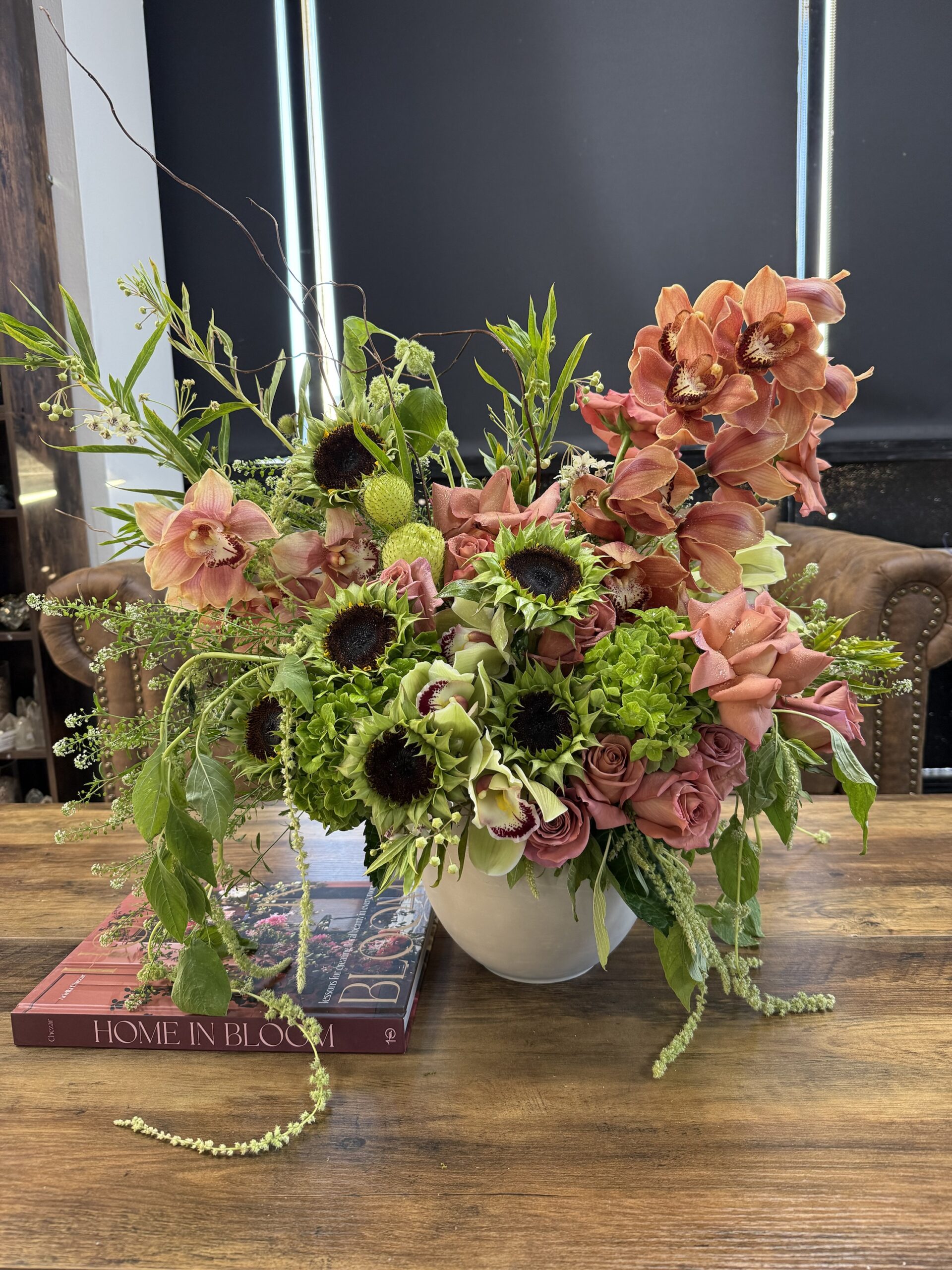 A vibrant arrangement of pink orchids, roses, green hydrangeas, and sunflowers with dark centers sits in the Not Ur Typical Gal Ceramic Pot on a wooden table next to books, one titled Home in Bloom.