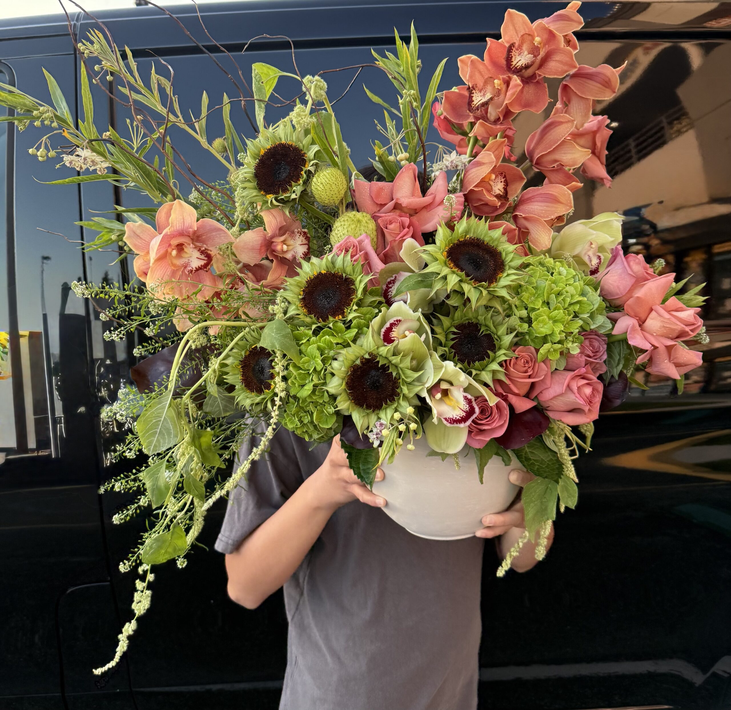 A person holds the Not Ur Typical Gal - Ceramic Pot, a vibrant floral arrangement with green hydrangeas, pink roses, orange orchids, sunflowers, and greenery; a black vehicle and buildings appear in the background.