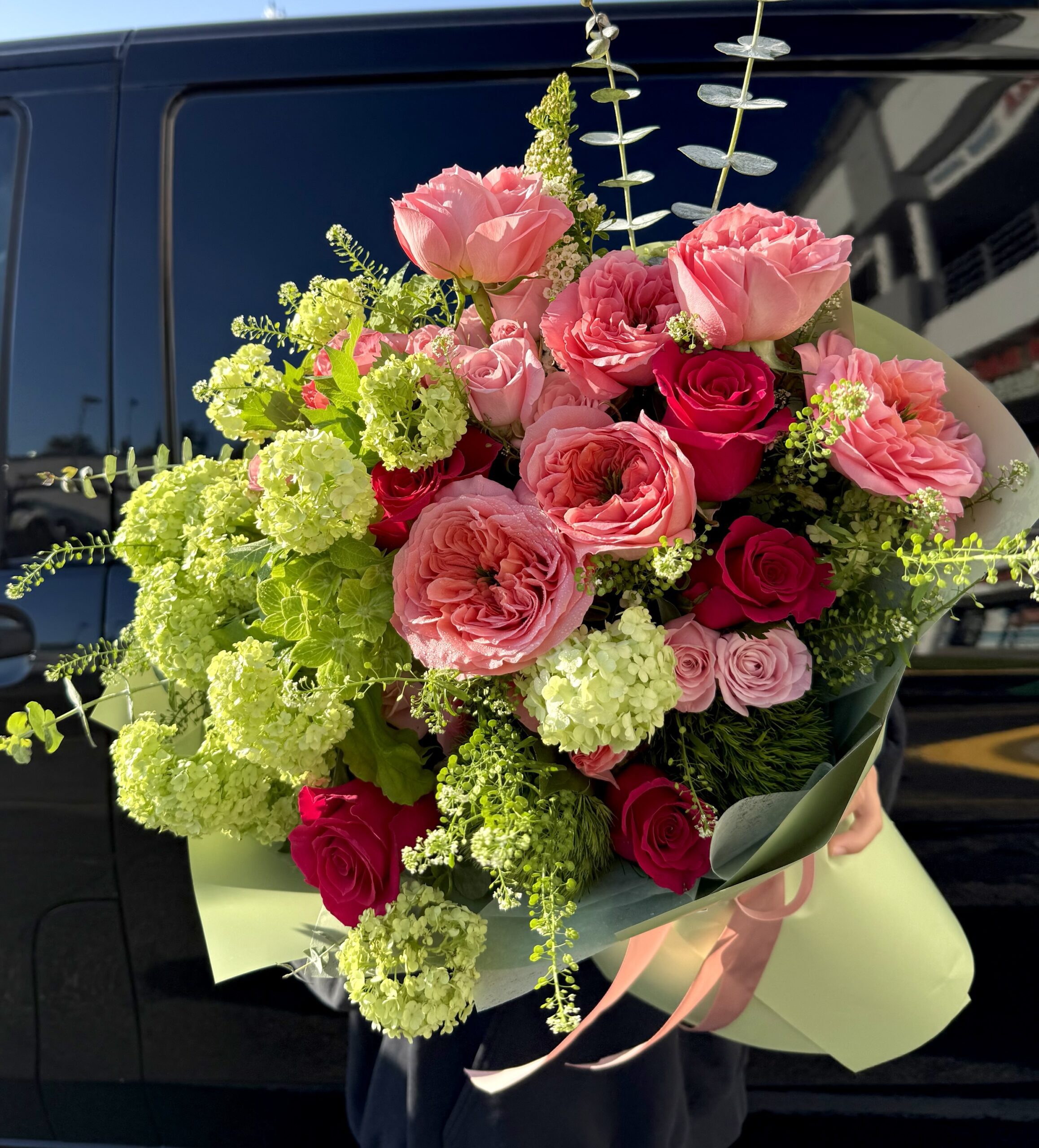 The Bon Voyage! - Wrapped Bouquet features pink roses, pink peonies, green hydrangeas, and assorted greenery, elegantly wrapped in light green paper with a pink ribbon.