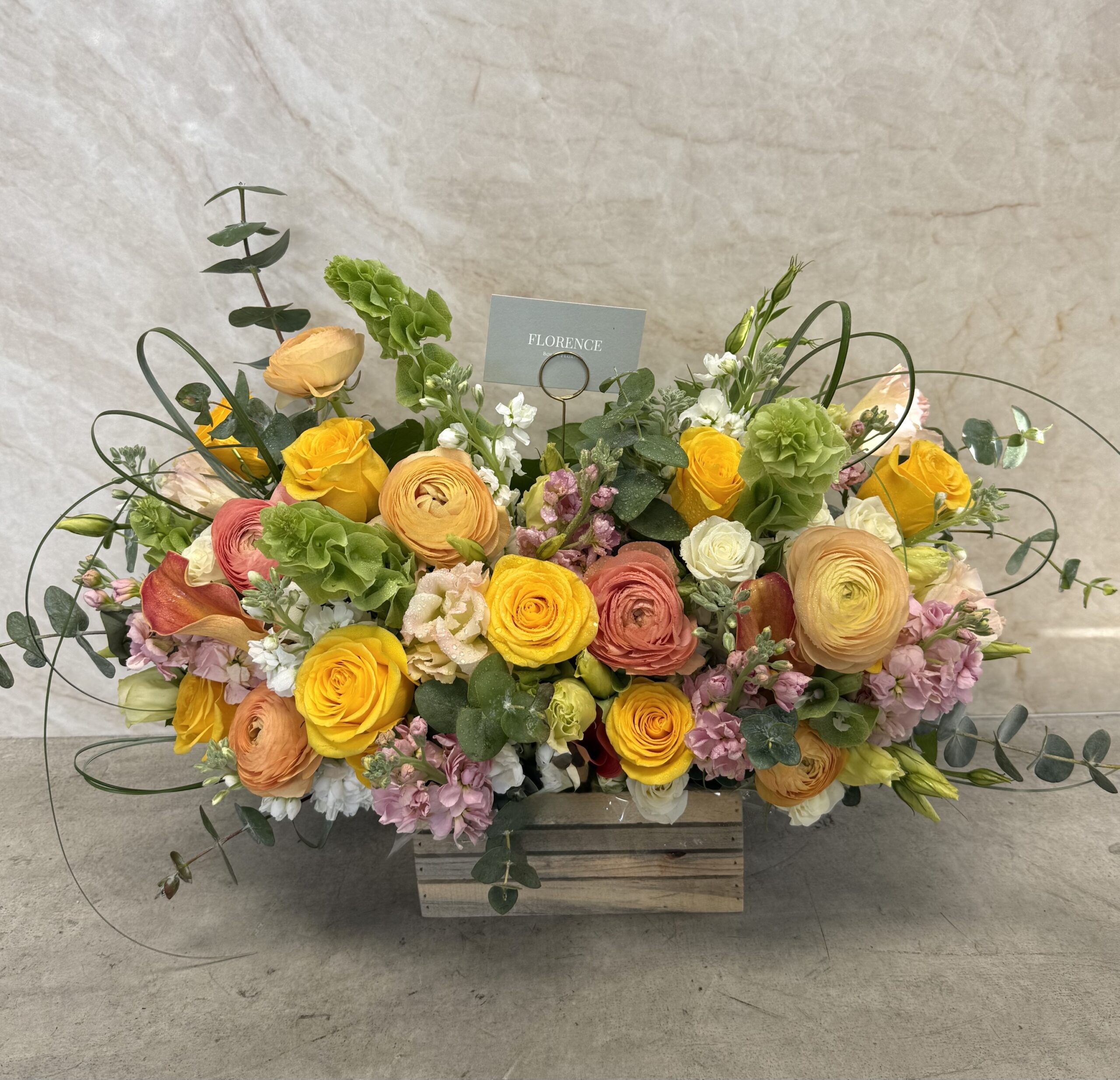 The Clementine Kisses - Wood Pot features a rectangular wooden box filled with yellow, orange, pink, and white flowers, green leaves, and eucalyptus branches, set against a light marble background.