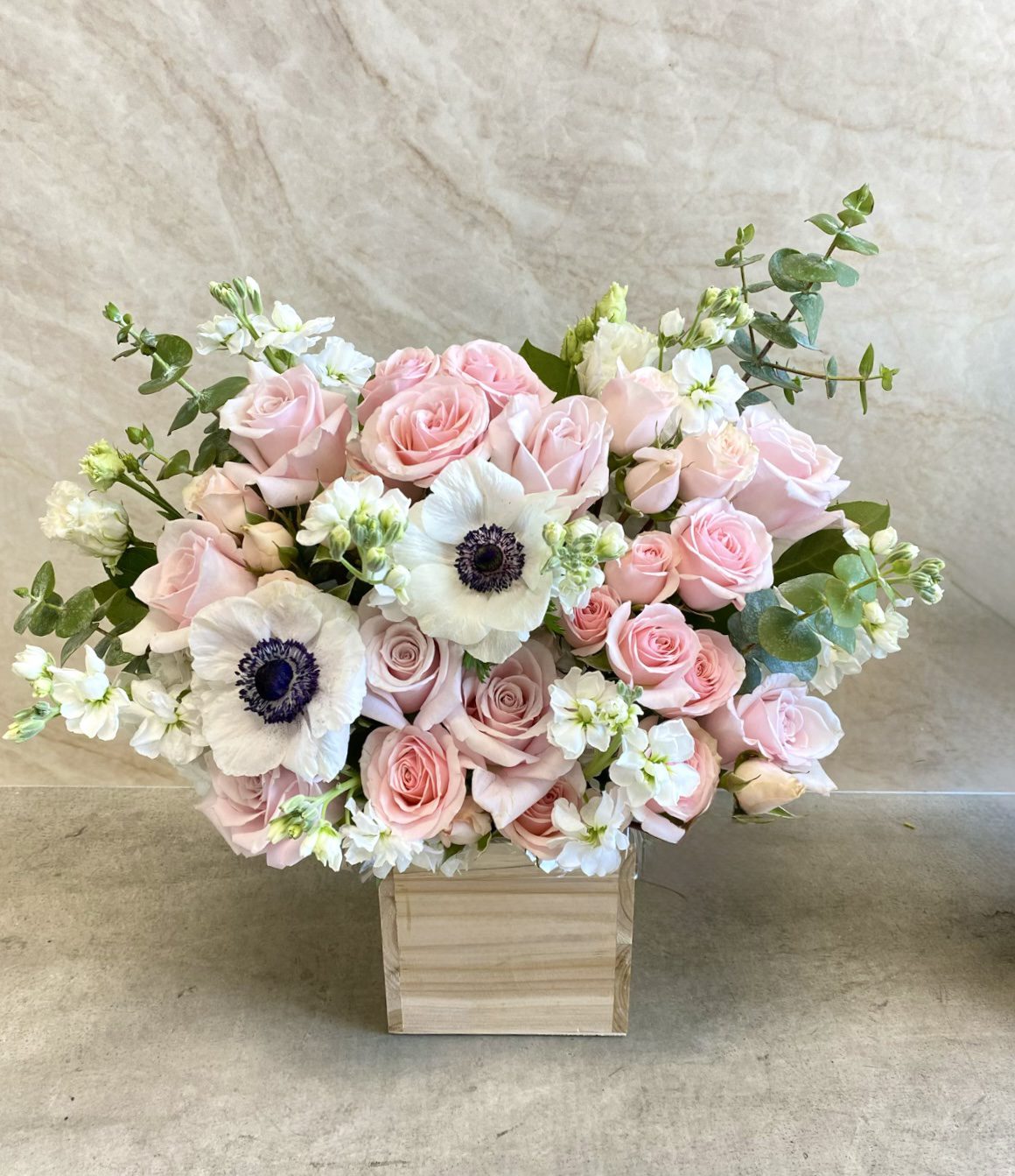 The Blush Hour - Wood Pot features light pink roses, white anemones with dark centers, small white blooms, and greenery arranged in a wooden vase against a light beige background.