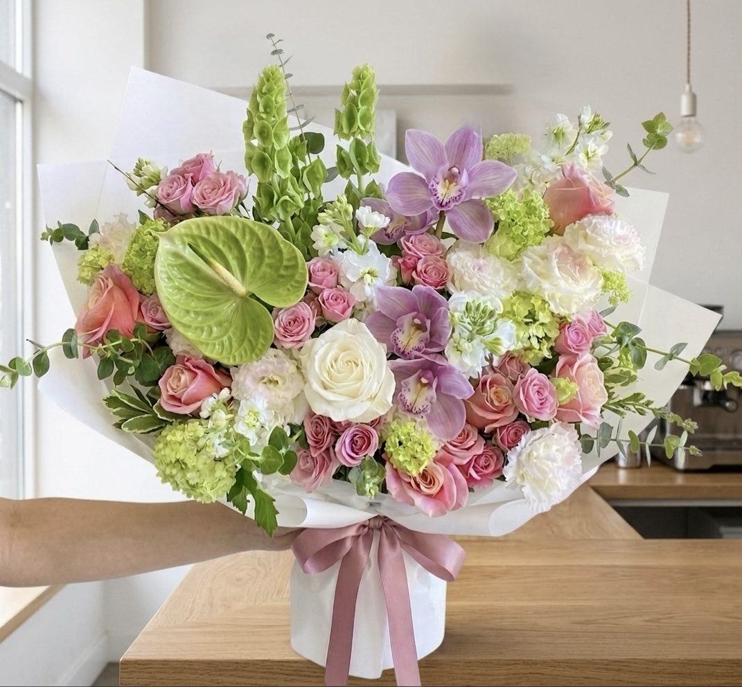 A hand displays the Fleur de Chérie - Wrapped Bouquet with pink, white, and purple flowers, green leaves, and anthurium, wrapped in white paper with a pink ribbon, shown in a modern kitchen.