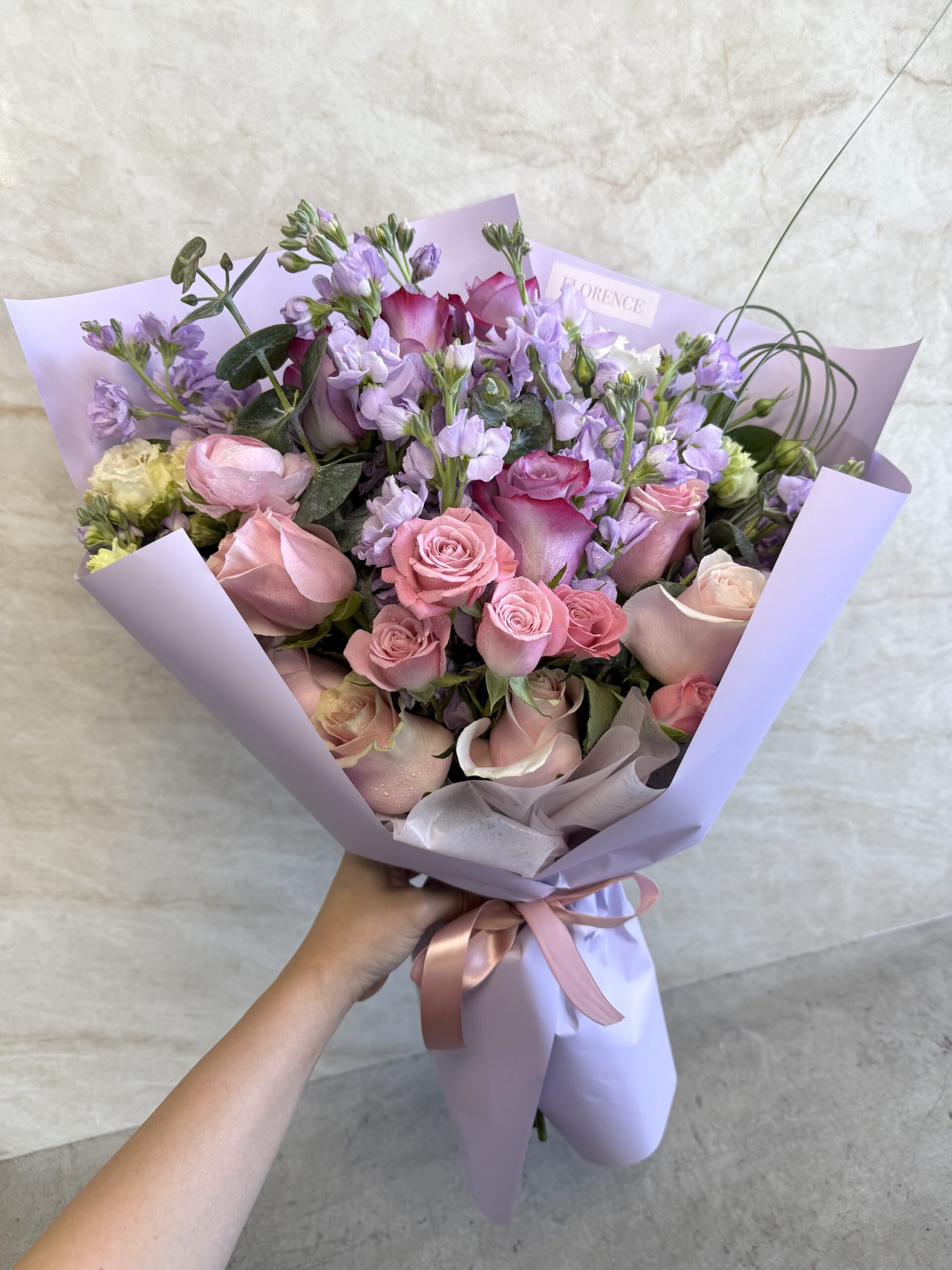 A hand holds the Bacio Dolce - Wrapped Bouquet, featuring pastel pink and purple flowers—roses and other blooms—wrapped in light purple paper and finished with a satin ribbon.