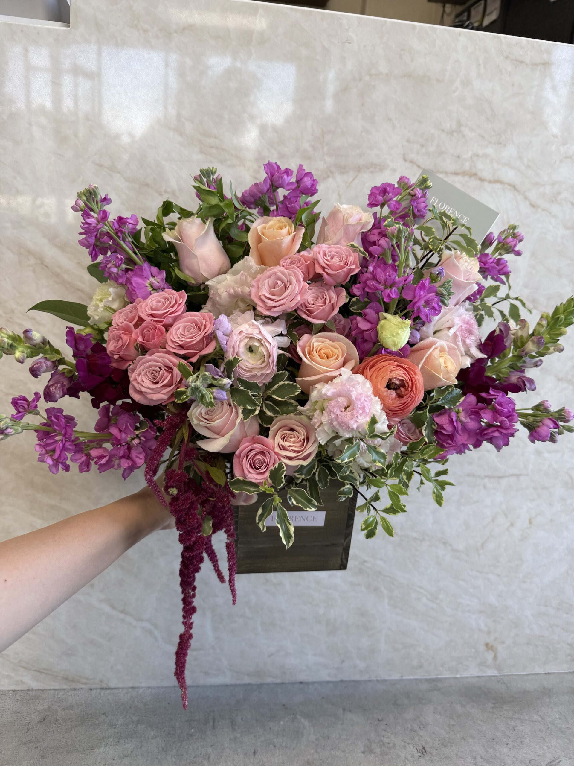A hand holds the Amore Mio - Wood Pot, a wooden box featuring a mixed bouquet of pink and peach roses, purple flowers, green foliage, and trailing red amaranthus against a light marble background.