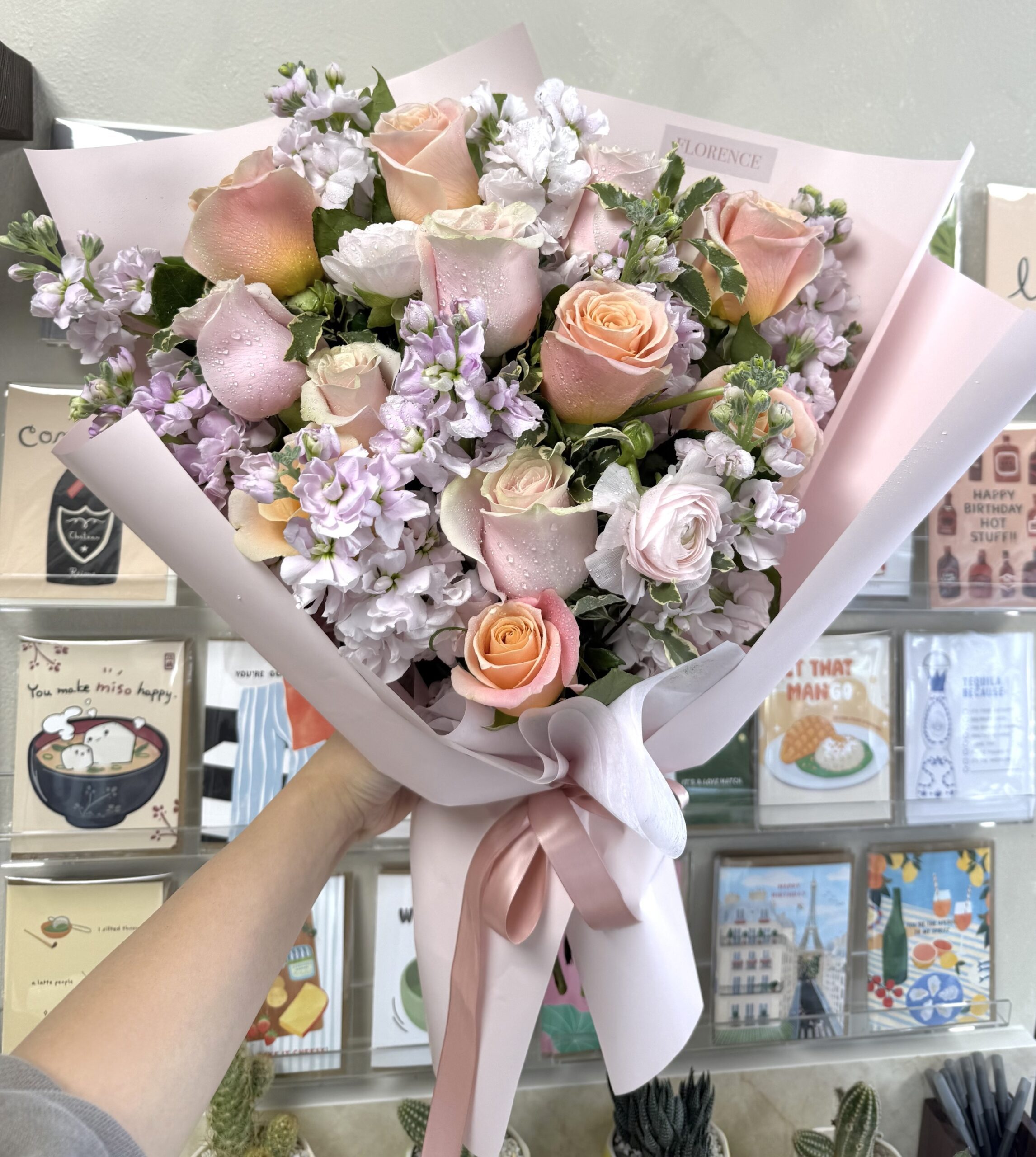 A hand holds the Jolie Bisous - Wrapped Bouquet, featuring pink and peach roses with light purple flowers in pink paper tied with a ribbon. Greeting cards are visible on shelves in the background.