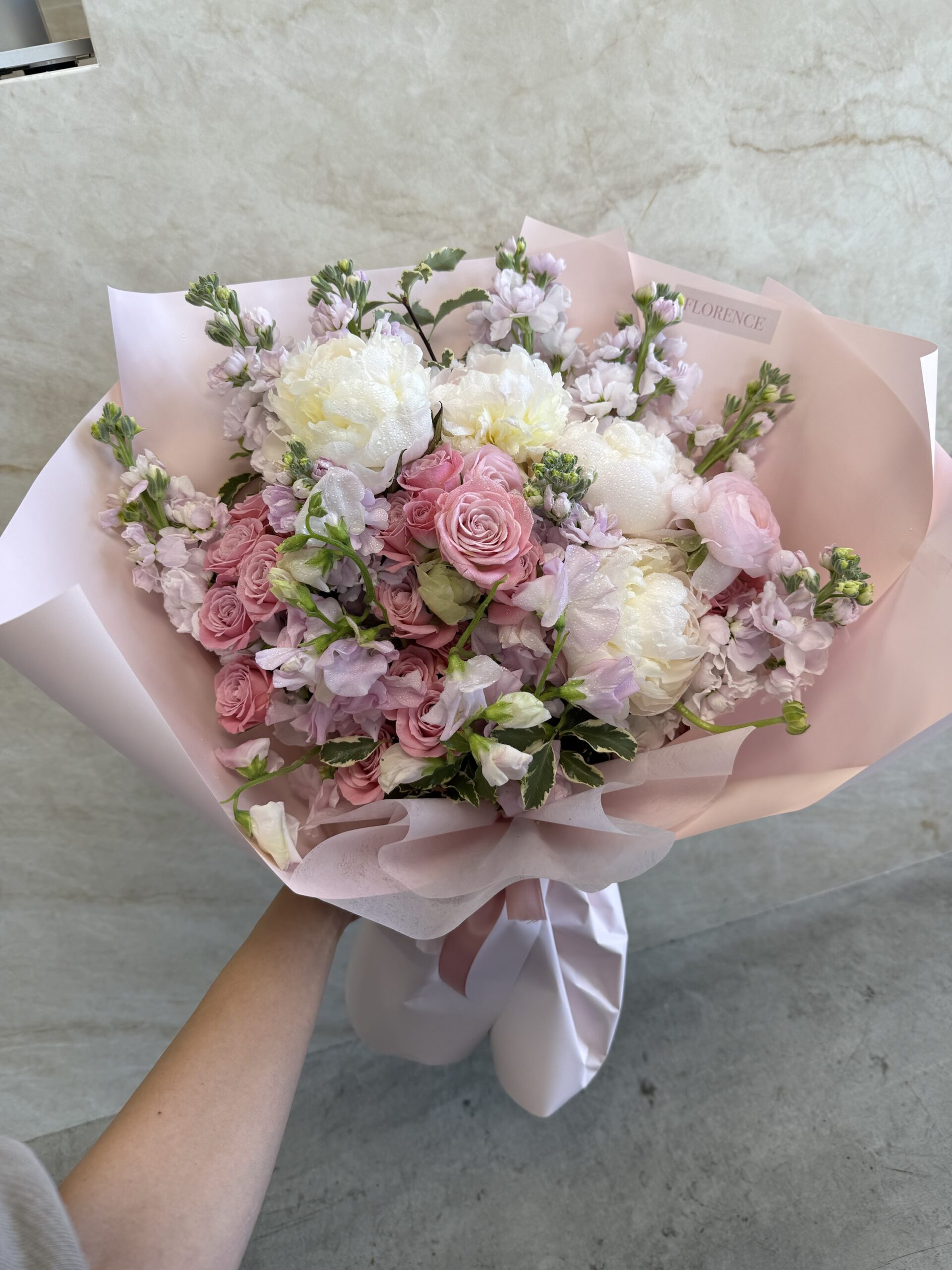 A hand holds the Fleur de Lune - Wrapped Bouquet, featuring pink and white roses, peonies, and delicate greenery wrapped in pale pink paper against a neutral backdrop.