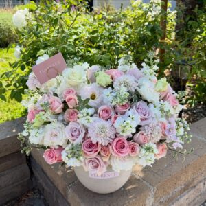 A lush bouquet of pink, white, and pale green roses with assorted flowers is arranged in the Maison de Flore - Ceramic Vase on a stone ledge outdoors, featuring a pink card marked FLORENCE nestled among the blooms.