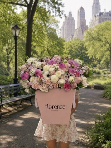 A person in a floral dress holds a large pink box of mixed flowers labeled “Blushing Beauty - Grand Flower Box,” standing on a path in a park with city buildings in the background.