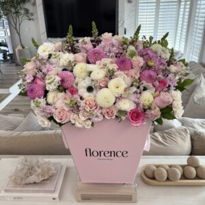 Large pink box overflowing with assorted pink, white, and purple flowers sits on a table in a modern living room. The box is labeled florence. Light streams in through white shutters behind the floral arrangement.