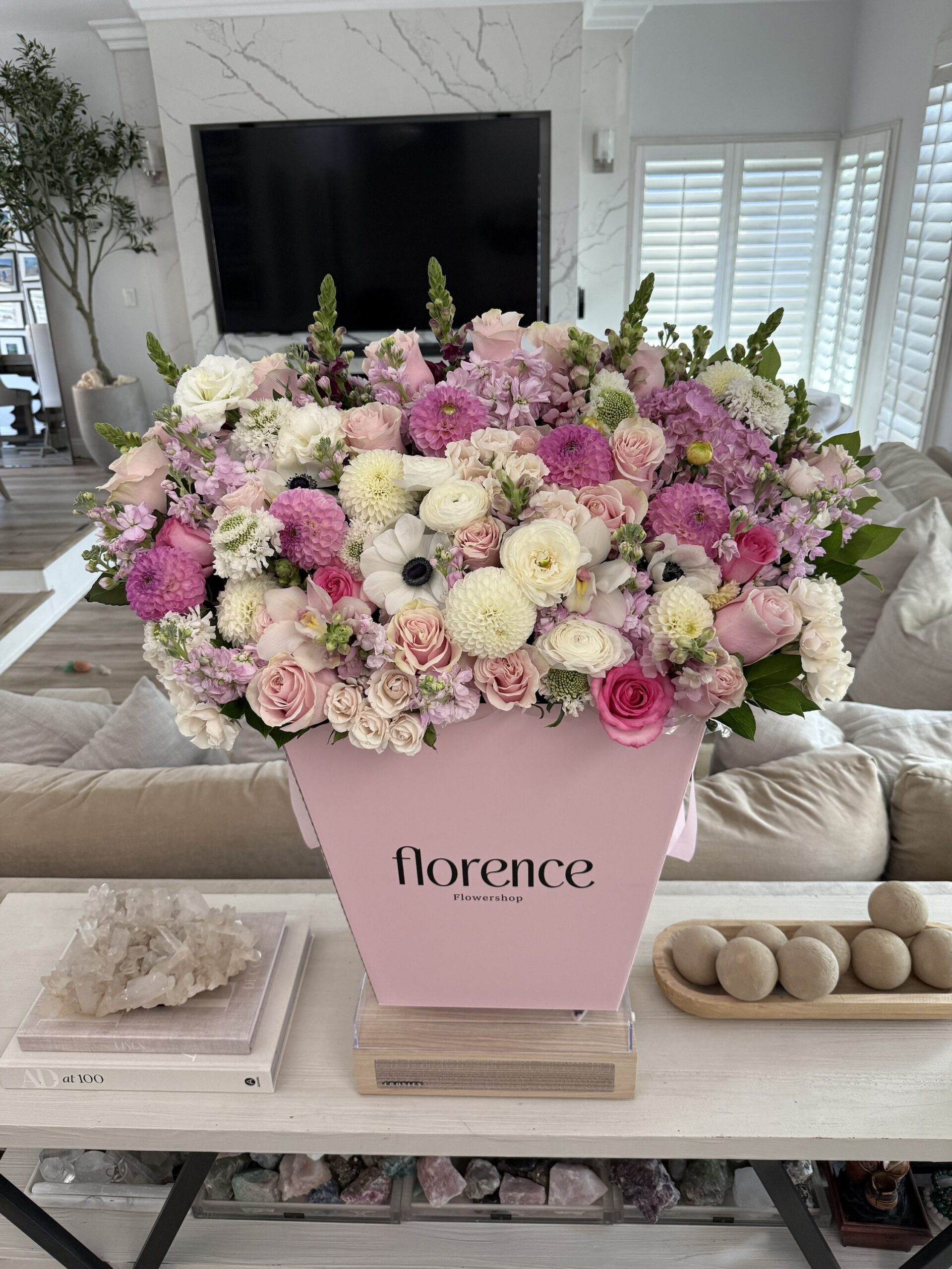 Large pink box overflowing with assorted pink, white, and purple flowers sits on a table in a modern living room. The box is labeled florence. Light streams in through white shutters behind the floral arrangement.