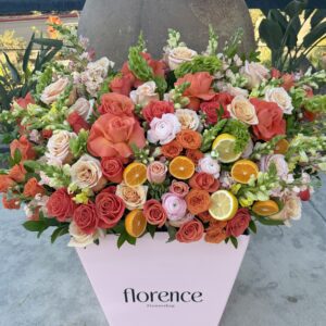 A large pink box labeled florence Flowershop holds a vibrant arrangement of orange, pink, and white roses, accented with fresh lemon and orange slices, set on a concrete surface outdoors.