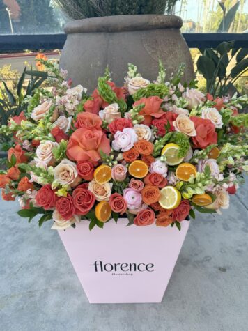 A large pink box labeled florence Flowershop holds a vibrant arrangement of orange, pink, and white roses, accented with fresh lemon and orange slices, set on a concrete surface outdoors.