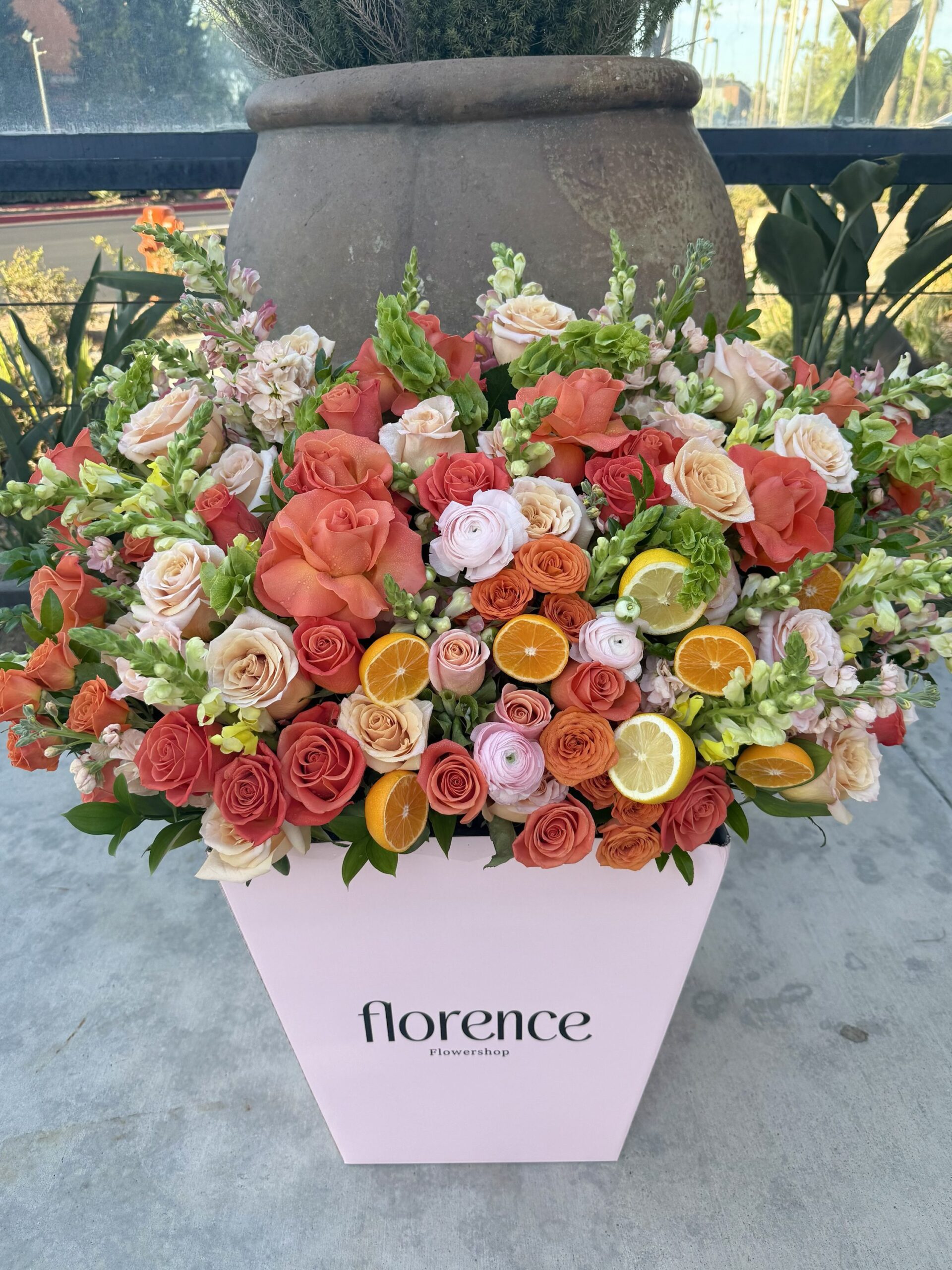 A large pink box labeled florence Flowershop holds a vibrant arrangement of orange, pink, and white roses, accented with fresh lemon and orange slices, set on a concrete surface outdoors.