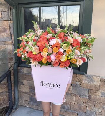 A person holds a large pink box labeled florence Flowershop filled with colorful roses, snapdragons, and citrus slices in front of a window against a stone wall.