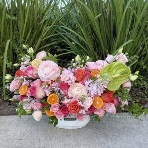 A vibrant floral arrangement with pink roses, ranunculus, lisianthus, green leaves, and orange citrus slices in a white container, set on a sidewalk with tall green plants in the background.