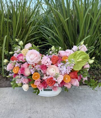 A vibrant floral arrangement with pink roses, ranunculus, lisianthus, green leaves, and orange citrus slices in a white container, set on a sidewalk with tall green plants in the background.