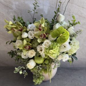 A floral arrangement in a vase featuring white roses, green orchids, hydrangeas, anthurium, greenery, and clusters of green grapes against a light stone background.