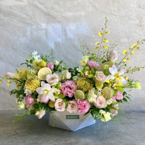 The Citrus & Champagne - Concrete Vase holds a vibrant floral arrangement featuring pink roses, lilies, yellow blooms, lush green leaves, and white accent flowers, all set against a light stone background.