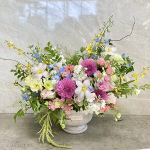 A large floral arrangement in a white vase features pink, purple, yellow, and white flowers with green foliage, set against a light marble background on a gray surface.