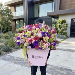 A person stands outside a modern house, holding the Hidden Gem - Grand Flower Box filled with vibrant, multicolored flowers. The person’s face is hidden behind the blooms.