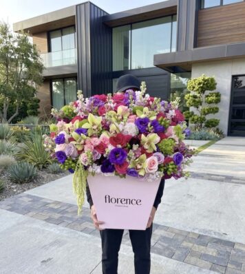 A person stands outside a modern house, holding the Hidden Gem - Grand Flower Box filled with vibrant, multicolored flowers. The person’s face is hidden behind the blooms.