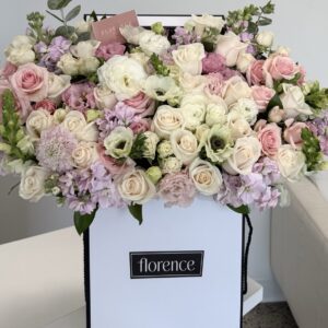 A white box labeled florence holds a large, lush arrangement of pastel roses, lisianthus, and other flowers in shades of white, pink, and lavender, displayed on a table near a white couch.