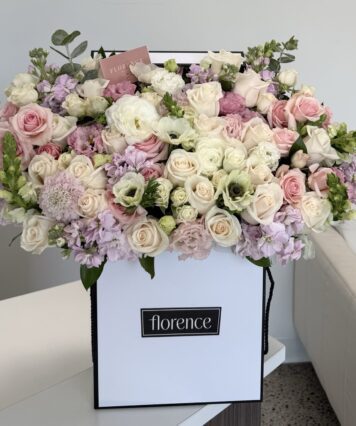 A white box labeled florence holds a large, lush arrangement of pastel roses, lisianthus, and other flowers in shades of white, pink, and lavender, displayed on a table near a white couch.