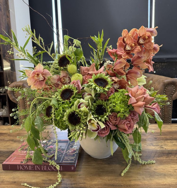 A vibrant arrangement of pink orchids, roses, green hydrangeas, and sunflowers with dark centers sits in the Not Ur Typical Gal Ceramic Pot on a wooden table next to books, one titled Home in Bloom.